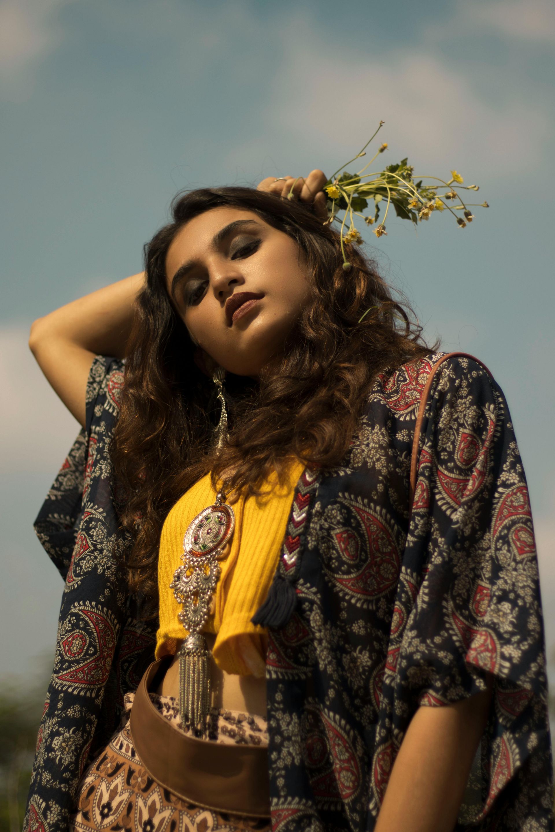 Woman in bohemian style with flowers, yellow top, paisley robe, against a blue sky.