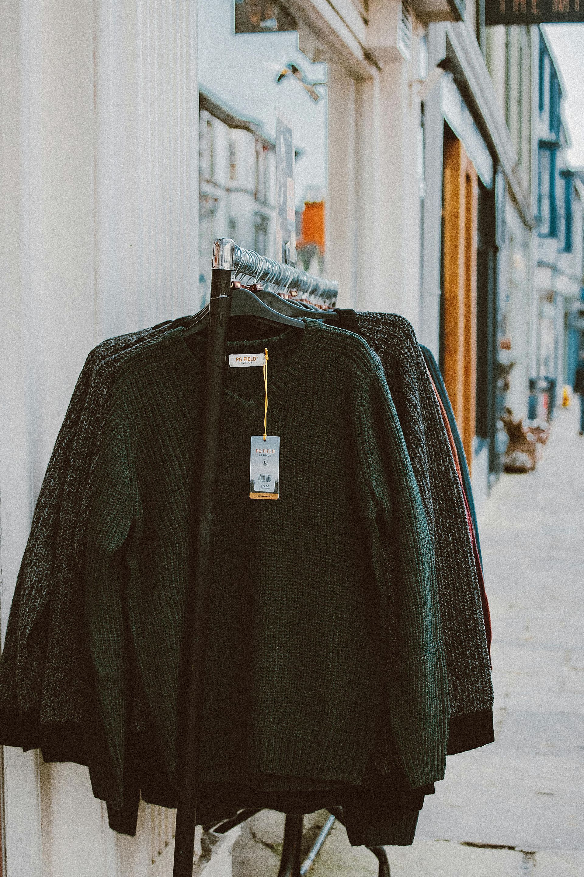 Dark green sweaters hanging on a rack outside a store on a street.