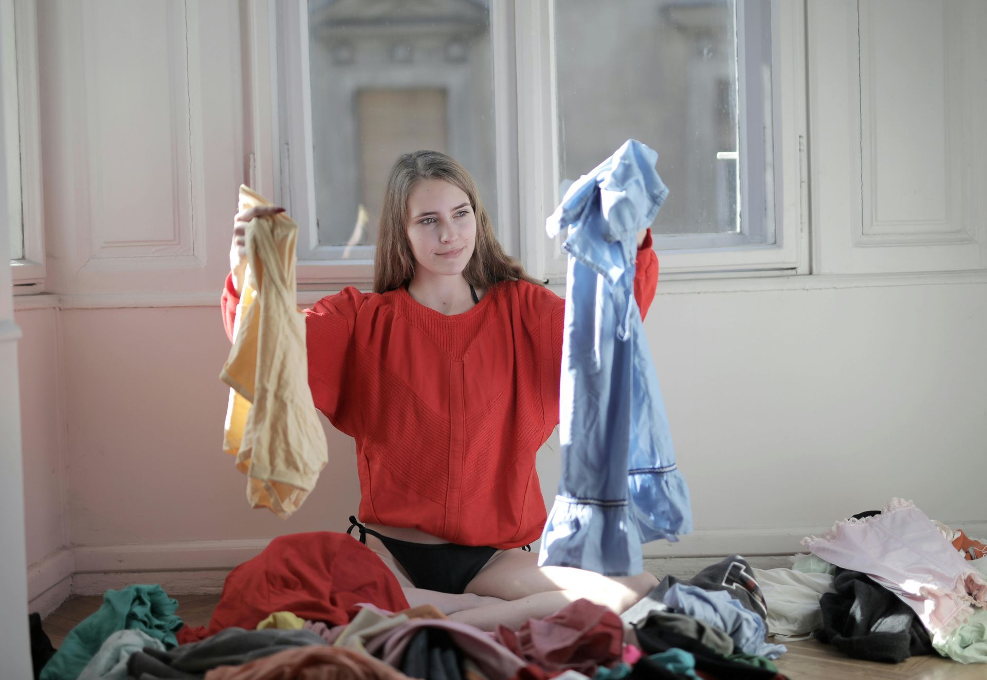 Woman in red sweater choosing between a yellow and blue top, surrounded by clothes on the floor near a window.