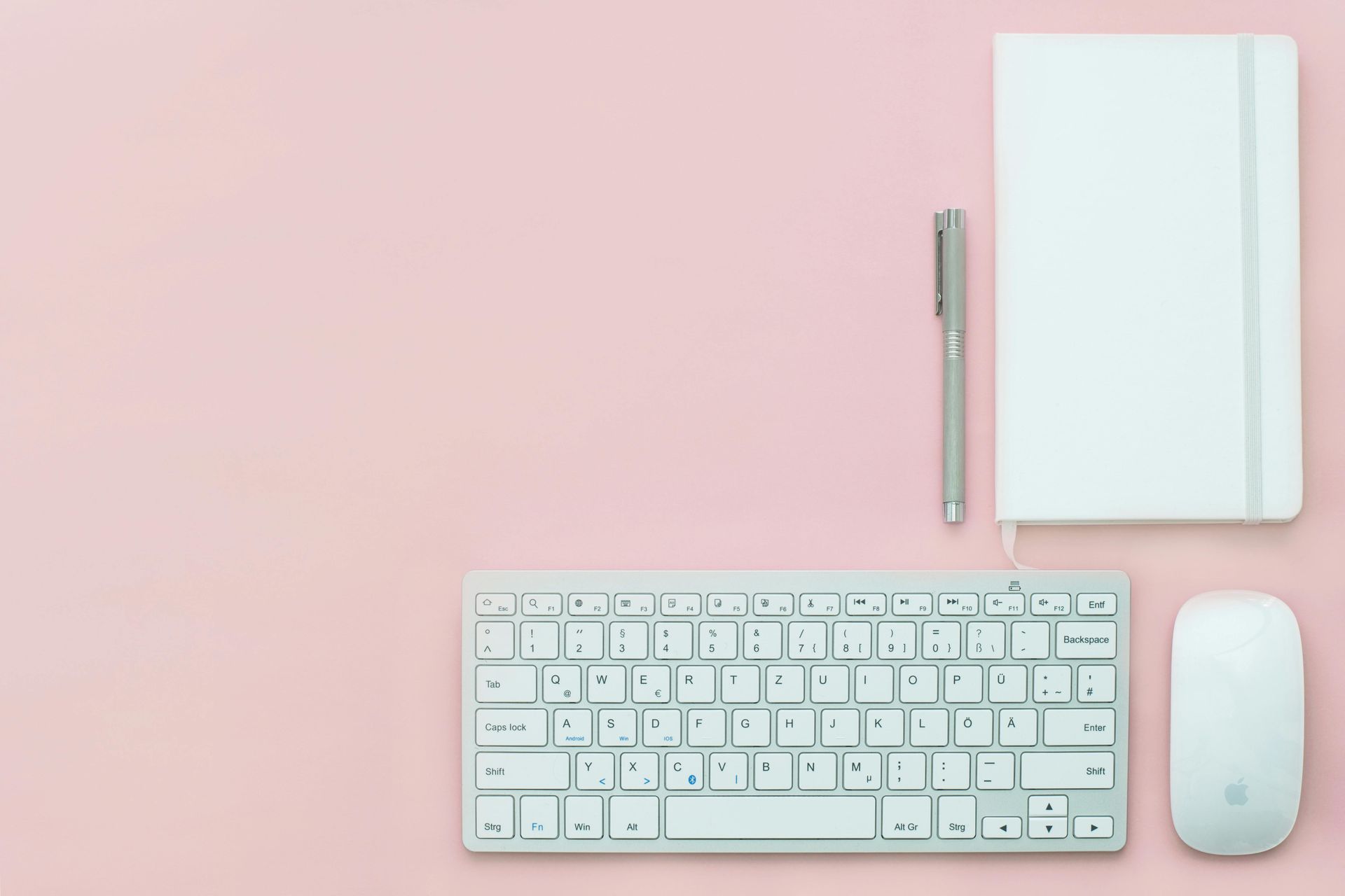 White keyboard, mouse, pen, and notebook on a pink background.