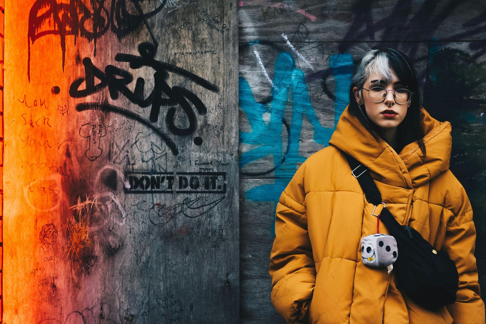 Woman in yellow coat with split-dye hair, standing in front of a graffiti-covered wall.