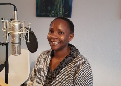 A woman smiling sits in front of a microphone in a studio. Grey sweater, light background. Swahili