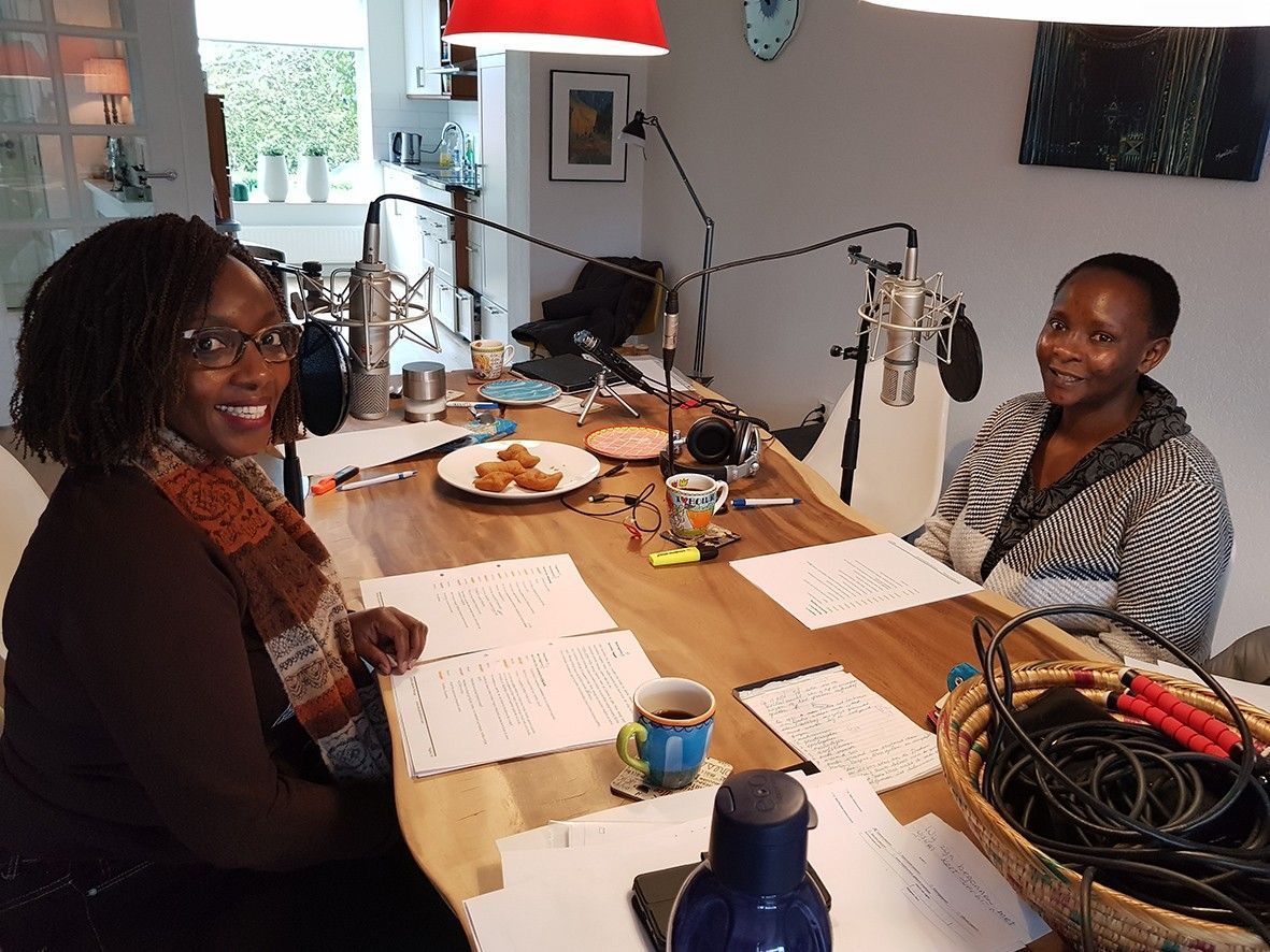 Two women at a table with microphones, papers, and snacks, likely recording a podcast in a home setting. Swahili