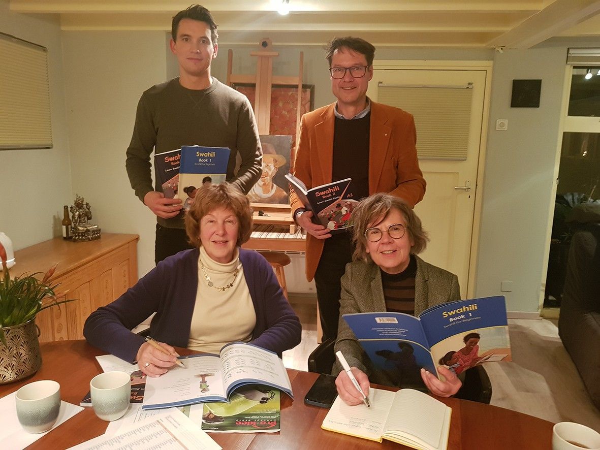 Four people holding books, possibly authors, gather indoors. Two at a table writing, two standing behind. Swahili Grammar books