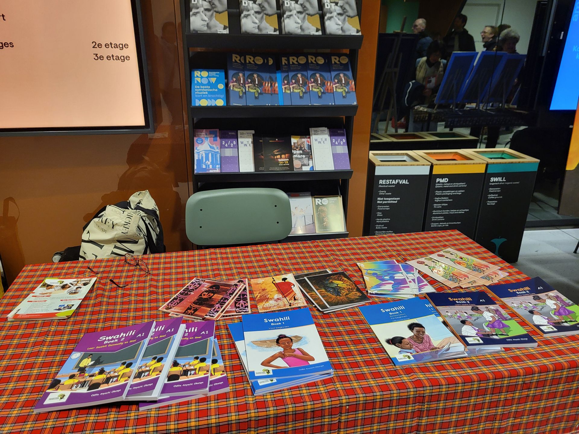 Books are displayed on a table with a patterned tablecloth. Shelves in the background hold more books. Swahili books
