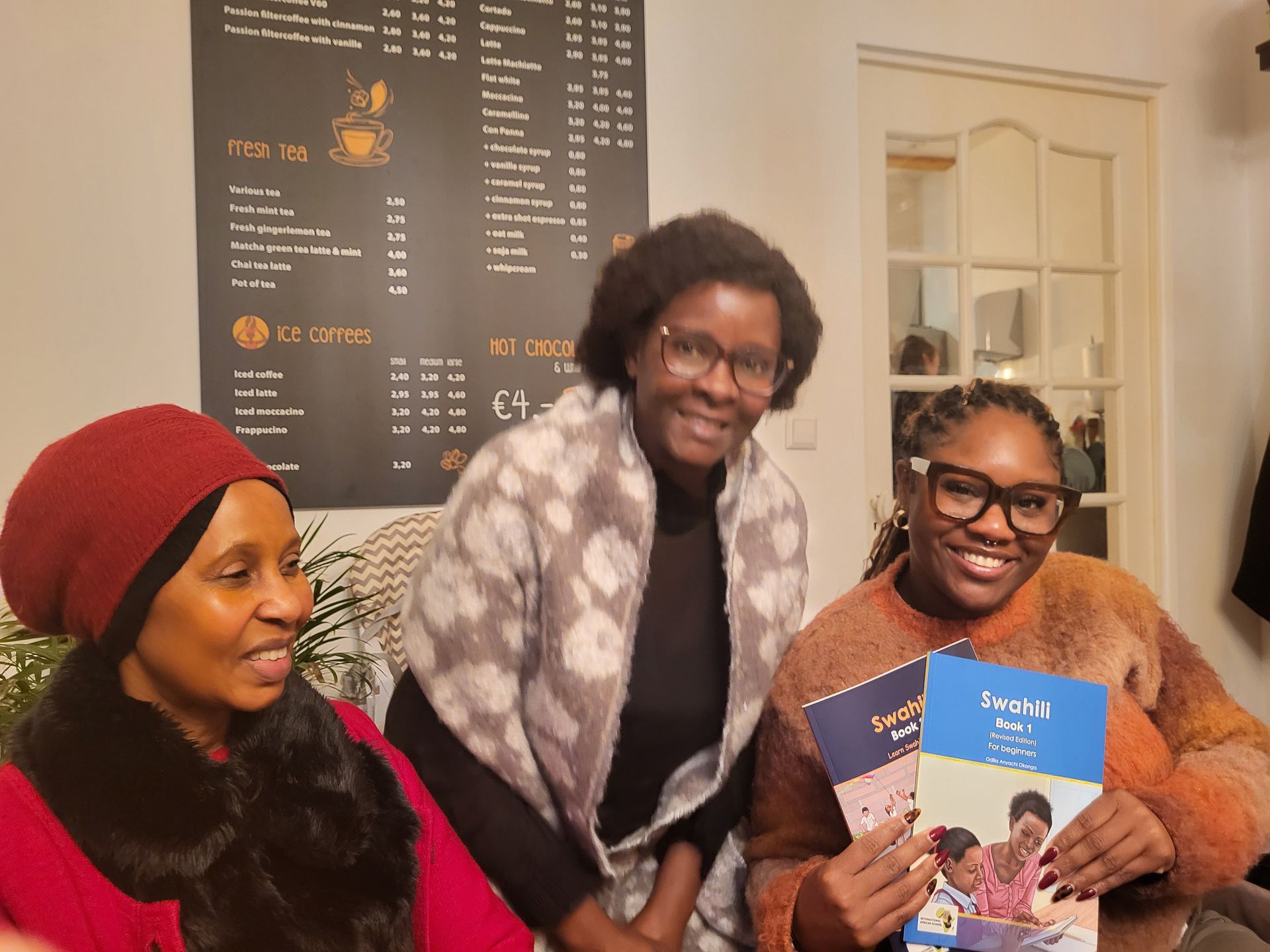 Three women smiling, holding booklets in a café setting with a menu on the wall.