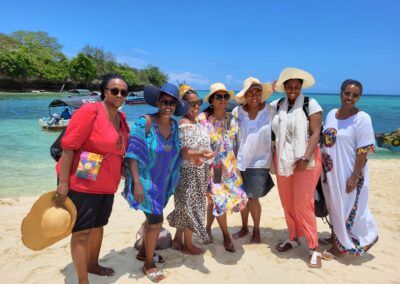 Seven people smiling on a sandy beach in Zanzibar near boats and blue water. They wear hats and light-colored clothing.. Educultural Travel