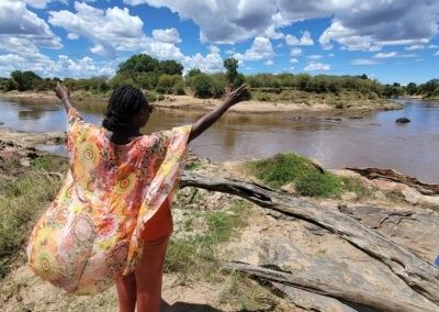 A person with arms raised stands by the Mara River, wearing a colorful shawl, on a sunny day.Educultural Travel