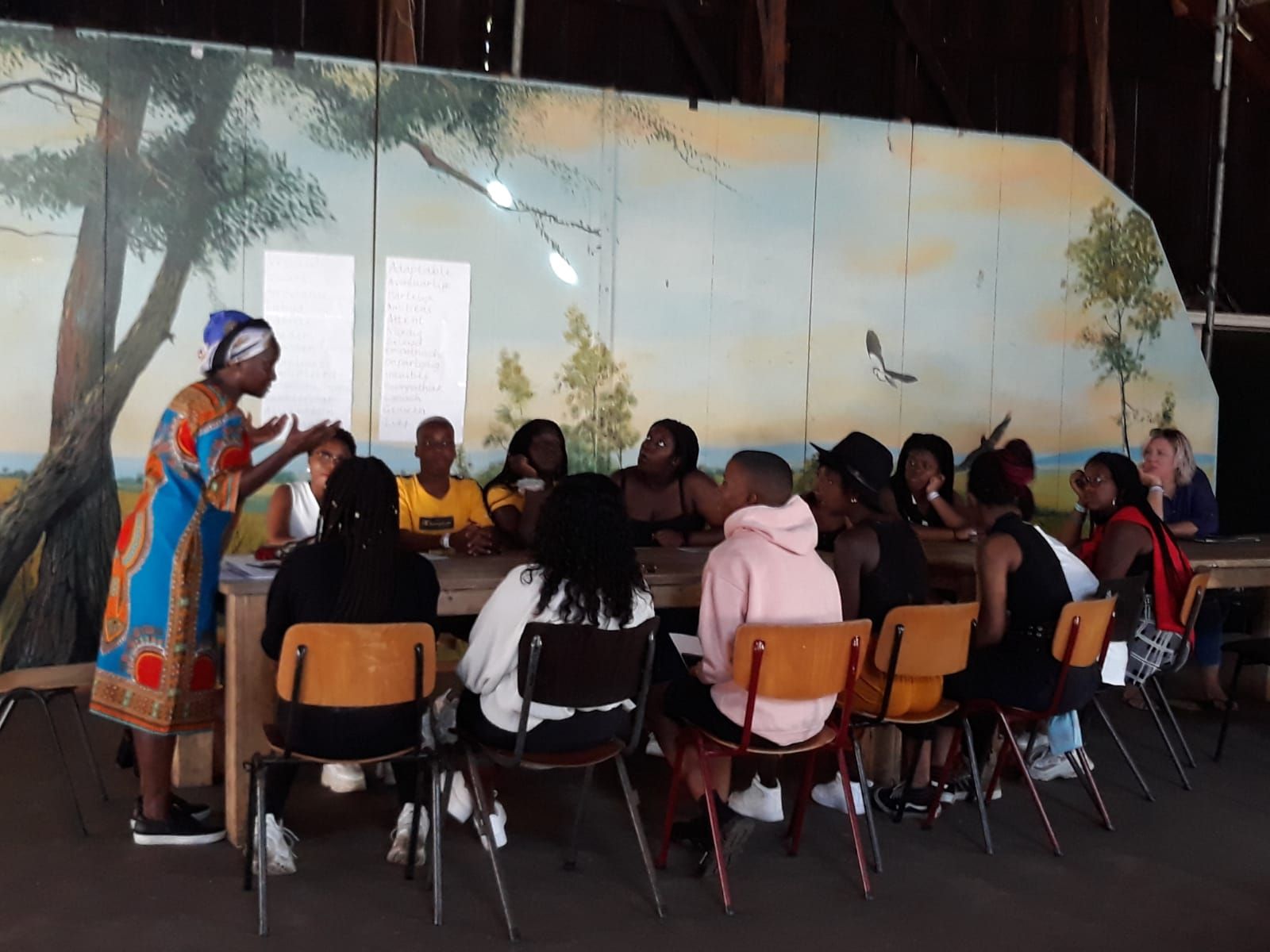 A woman in a colorful outfit speaks to a group seated at a long table against a painted backdrop.  Discovering Africa in the classroom