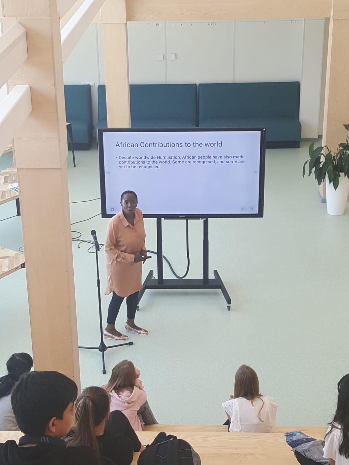 A woman is presenting in front of students, using a large screen and a microphone. School setting, wooden steps, light colors.  Discovering Africa in the classroom