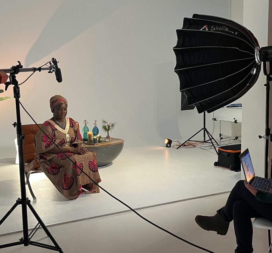 A woman in a patterned dress sits on a chair in a studio, illuminated by a softbox and lights. Someone works on a laptop.