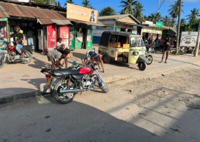 Motorcycles and a tuk-tuk on a dusty road in front of shops. People are gathered near the vehicles. Watamu Kenya. Educultural Travel