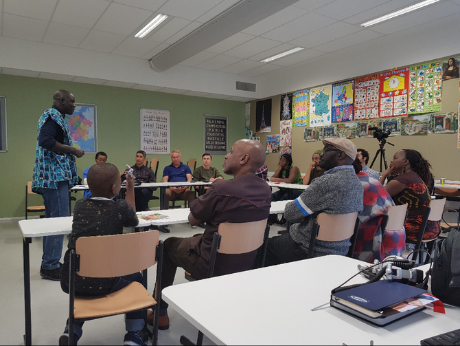 People in a classroom, with one person presenting. White tables, green wall, posters on the back wall.