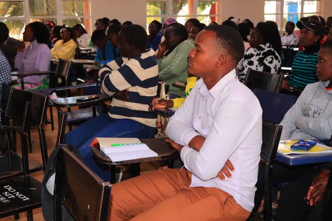 People in a classroom, listening to a lecture. Some are taking notes, all are facing the front.