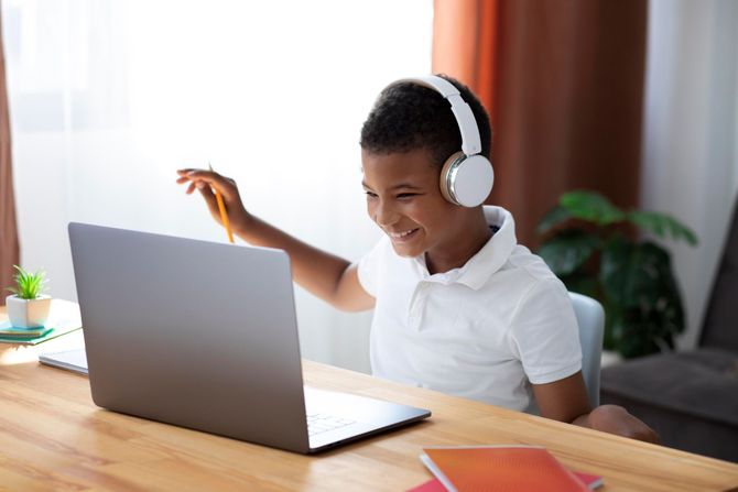 Boy wearing headphones smiles at a laptop, holding a pencil, in a well-lit room.