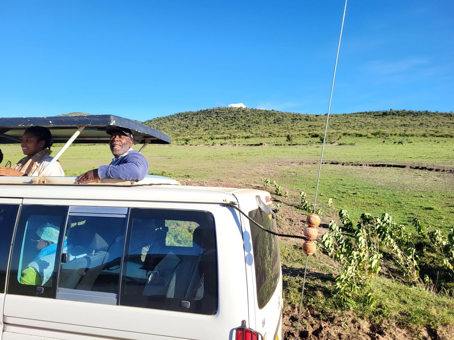 People in an open-top vehicle, scenic landscape. Safari. A white van with an antenna and people inside looking at a green hill. Educultural Travel. Safari