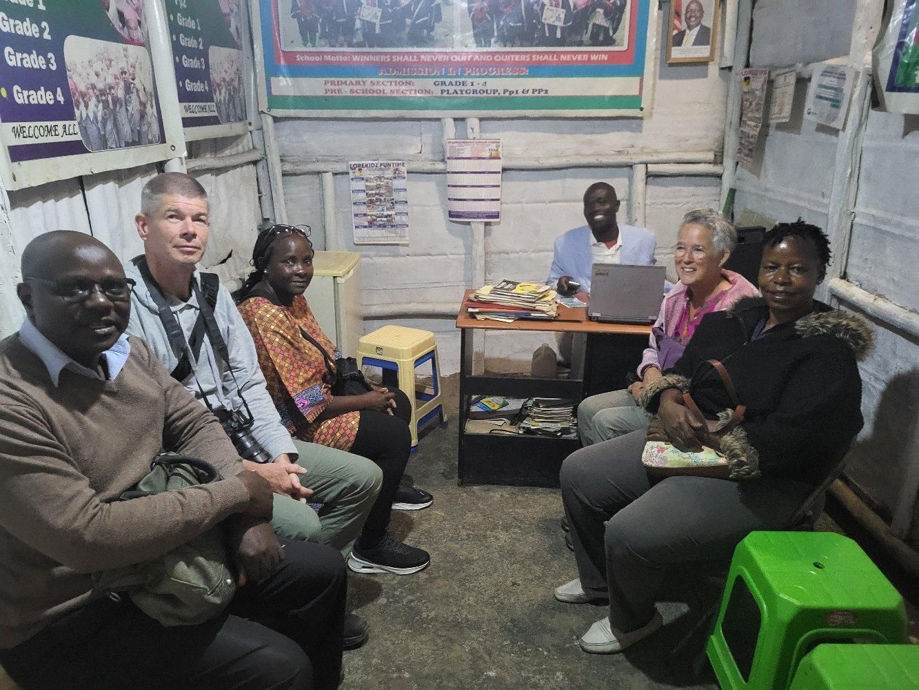 Group of people sitting in a small room, some facing the camera, next to a table with a laptop.