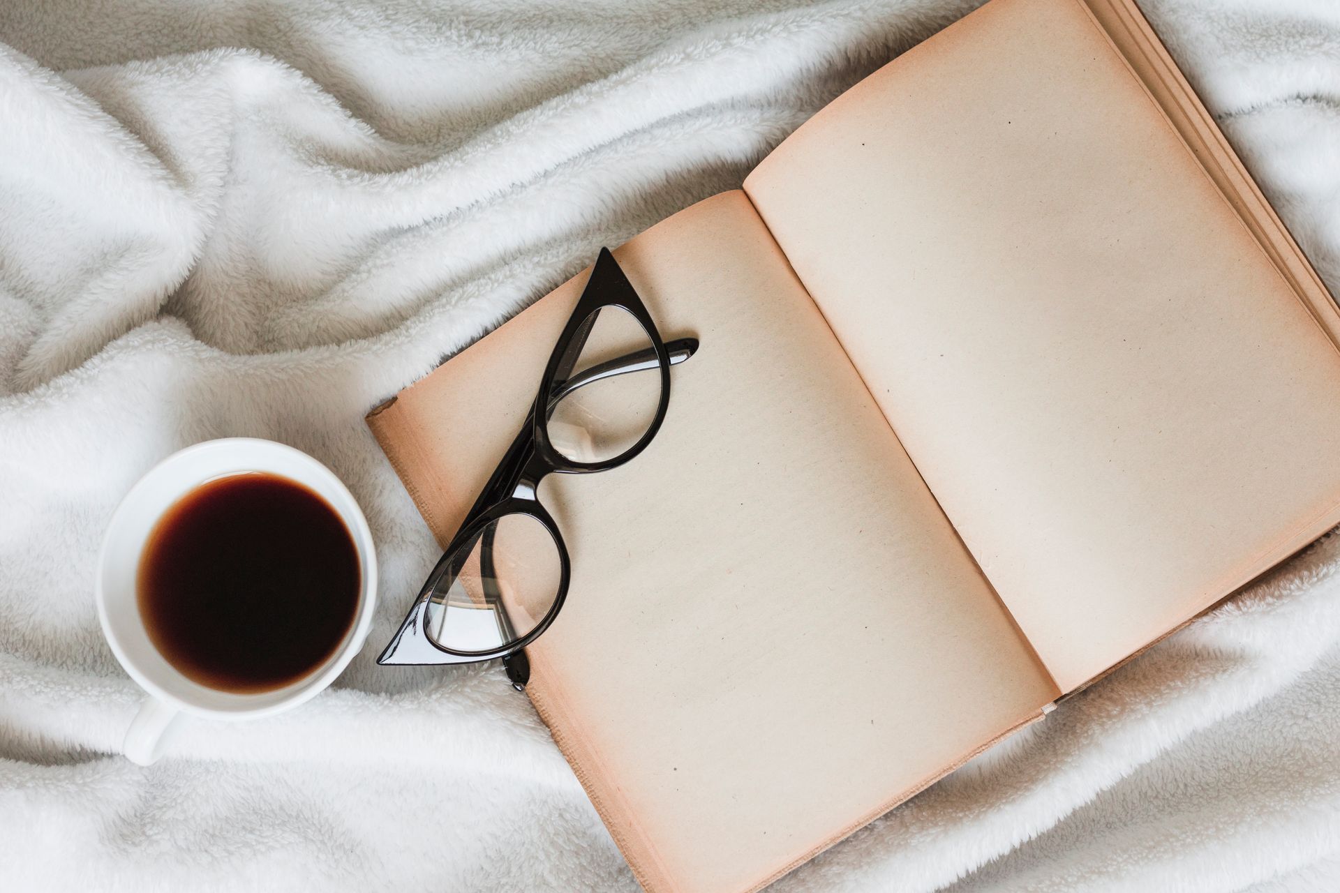 Open book, eyeglasses, and a coffee cup on a white blanket.