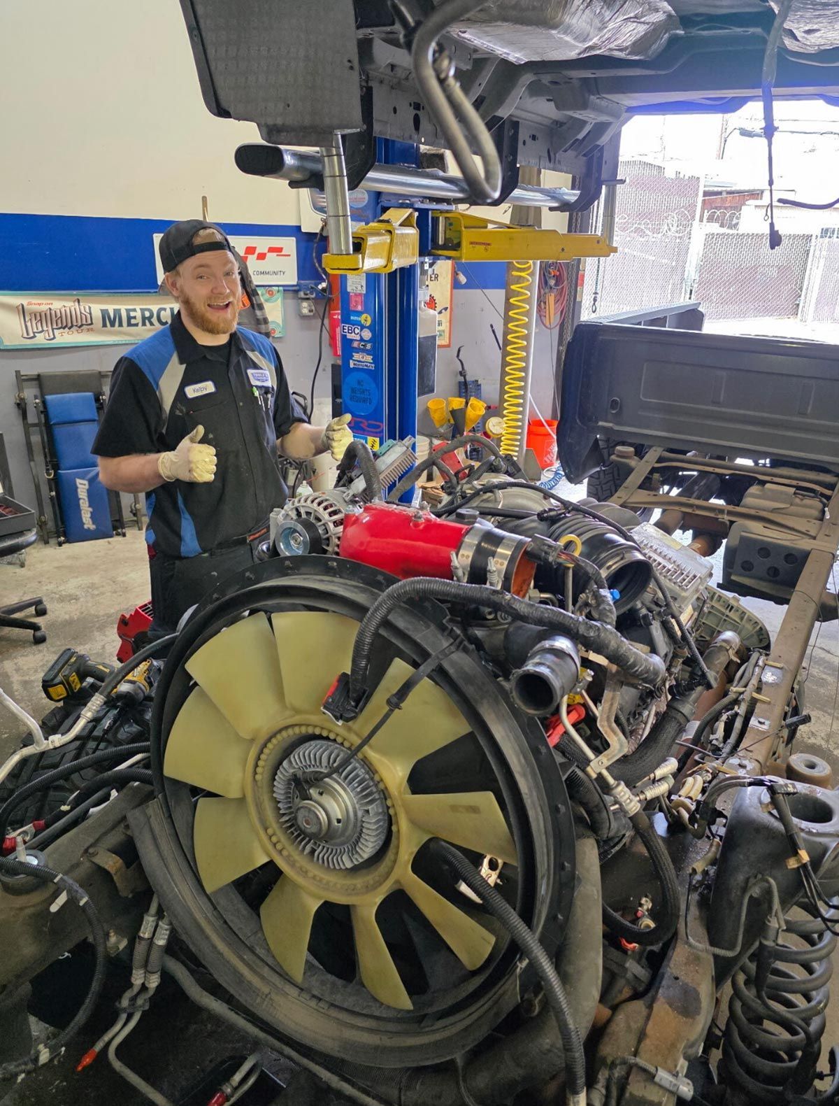 Mechanic beside vehicle engine in a shop giving thumbs up. Red parts, black hat, blue and black uniform.