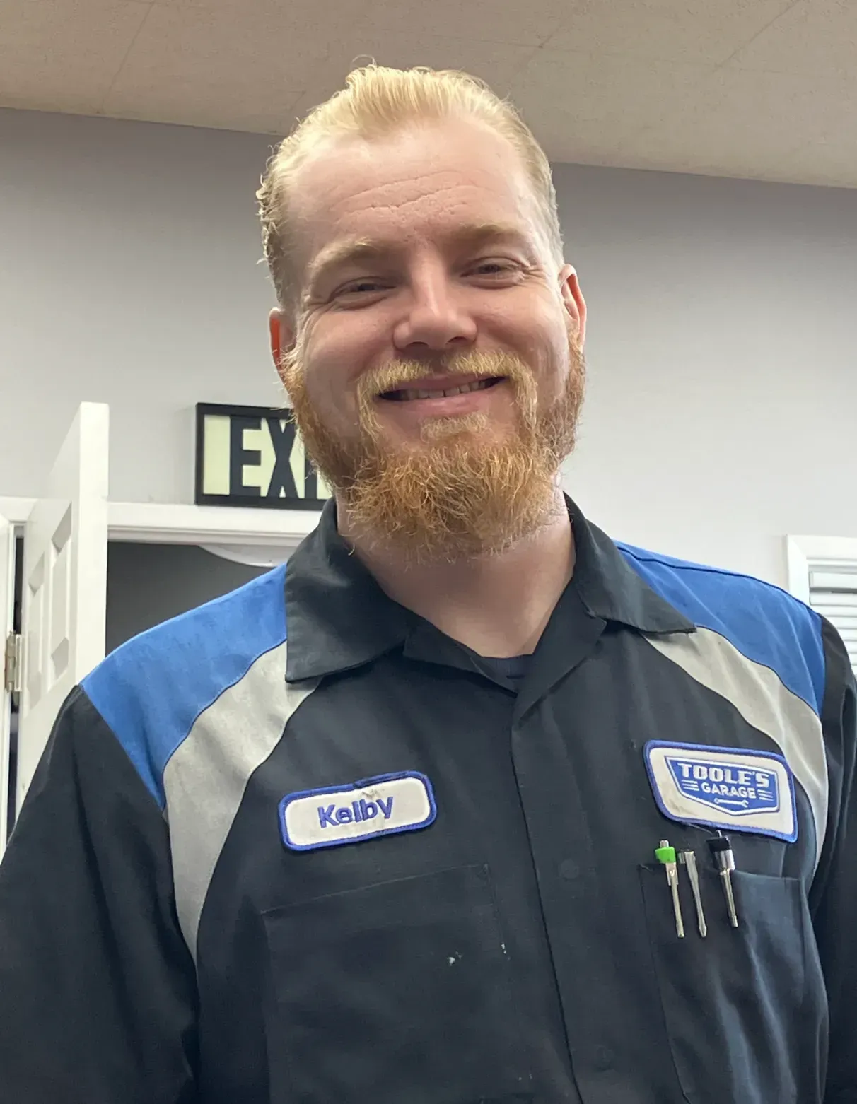 Man with blonde hair and beard in a blue and black uniform smiles in a shop.