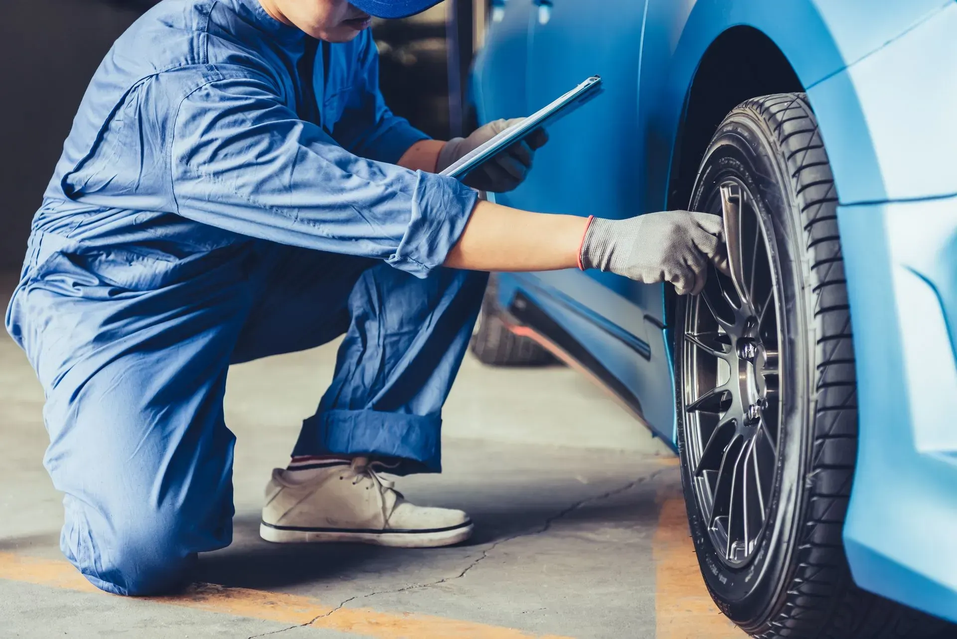 Mechanic in blue jumpsuit examining car tire with a tablet in a garage.