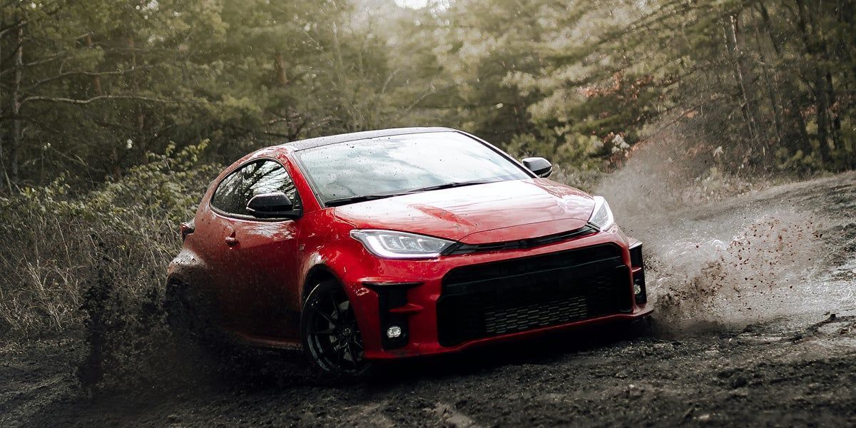 Red car driving through muddy terrain, spraying water. Surrounded by trees, overcast sky.