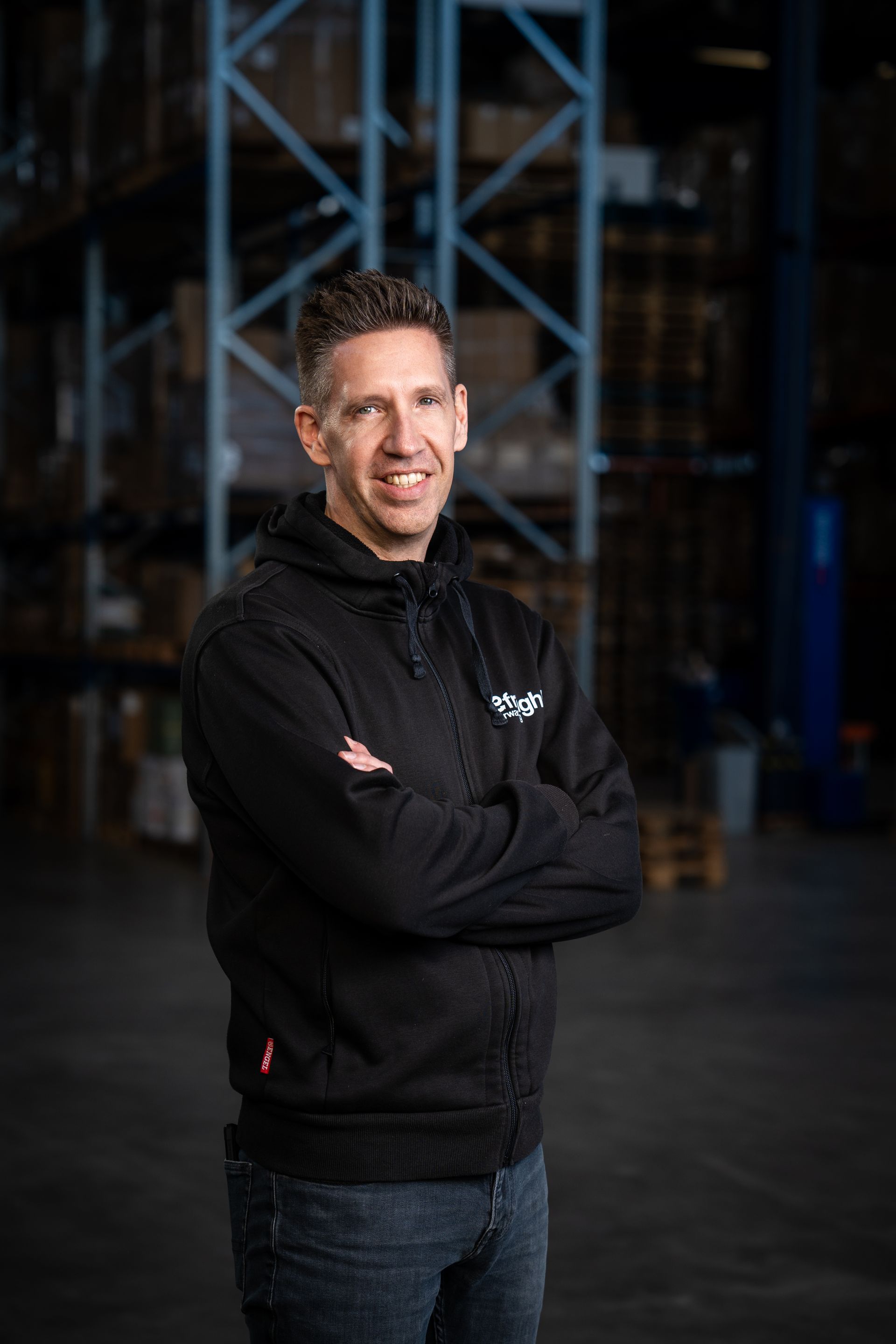 een man in een zwart jasje staat met zijn armen over elkaar in een pakhuis.