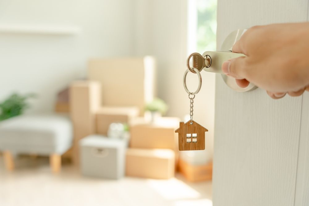 A hand opens a door with a house-shaped keychain, revealing a room filled with cardboard boxes, signifying moving house. — Diamond Real Estate QLD in Mango Hill, QLD