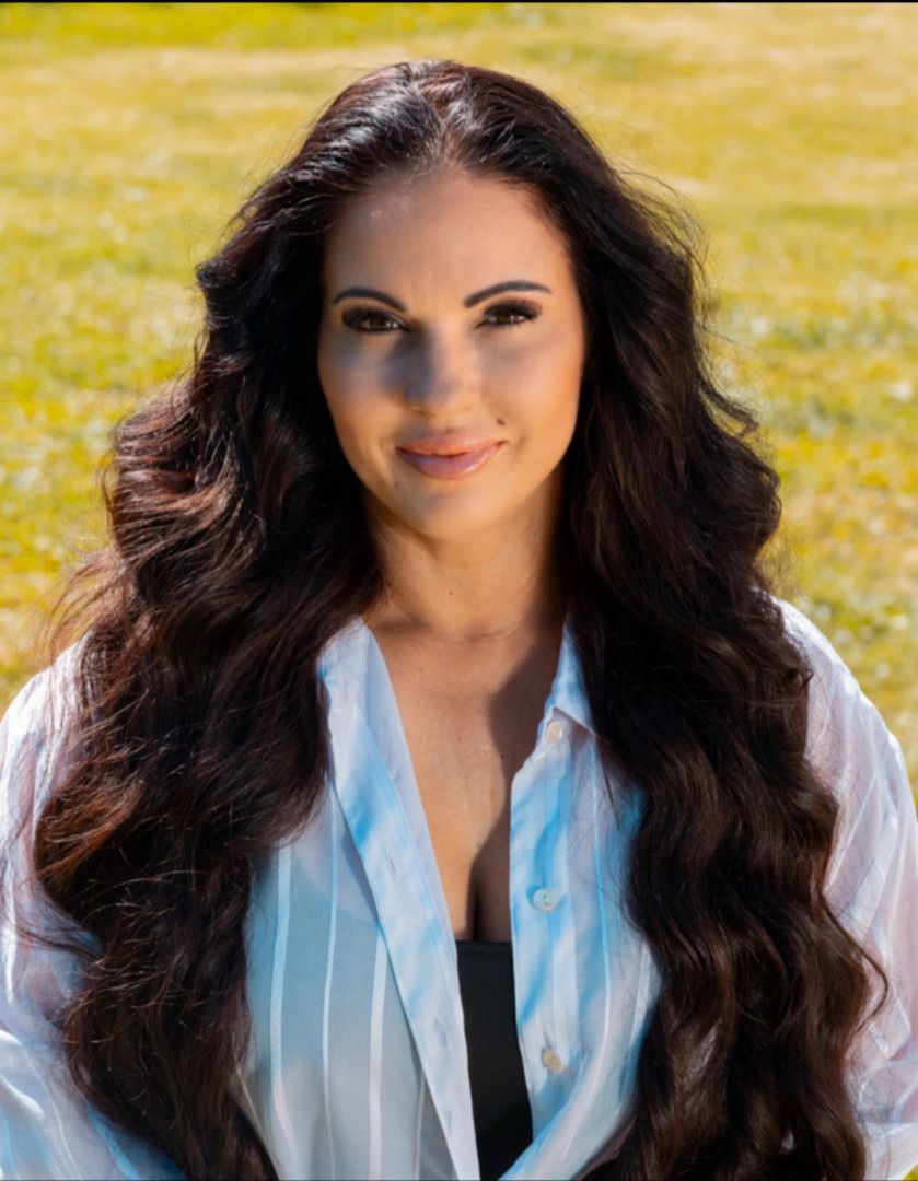 A woman with long, dark, wavy hair smiles against a blurred, sunlit grassy background, wearing a light-coloured shirt.