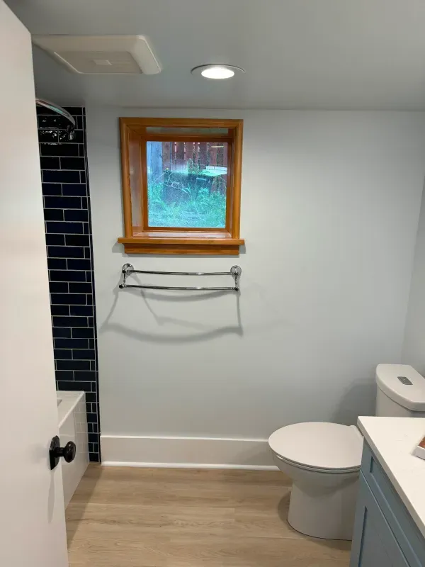 Bathroom with blue-tiled shower, light blue walls, wooden window frame, toilet, and silver towel rack.