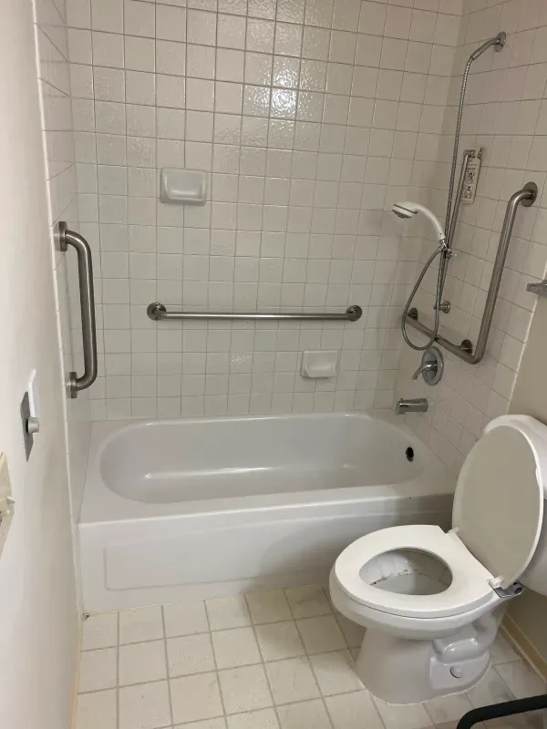 Accessible bathroom with tub, toilet, grab bars, and handheld shower. White tile throughout.