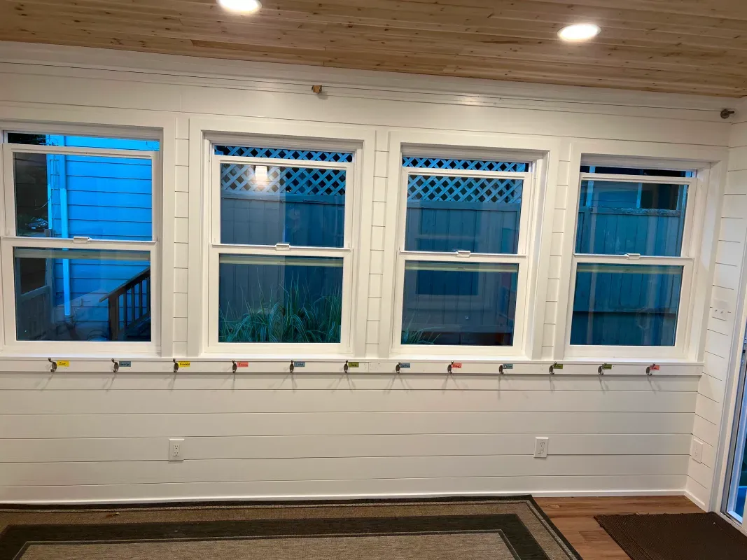 White-walled room with four windows, brown rug, and wooden ceiling. Windows look out onto a backyard.