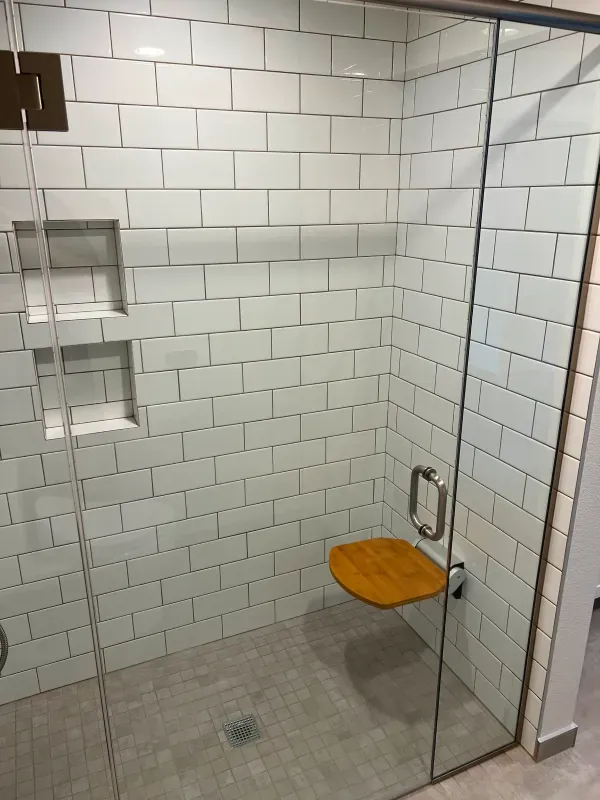 A modern bathroom shower with white brick tiles, glass doors, a wooden seat, and gray floor.