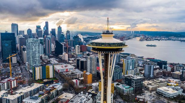 Seattle skyline featuring the Space Needle in foreground, with buildings, harbor, and cloudy sky.
