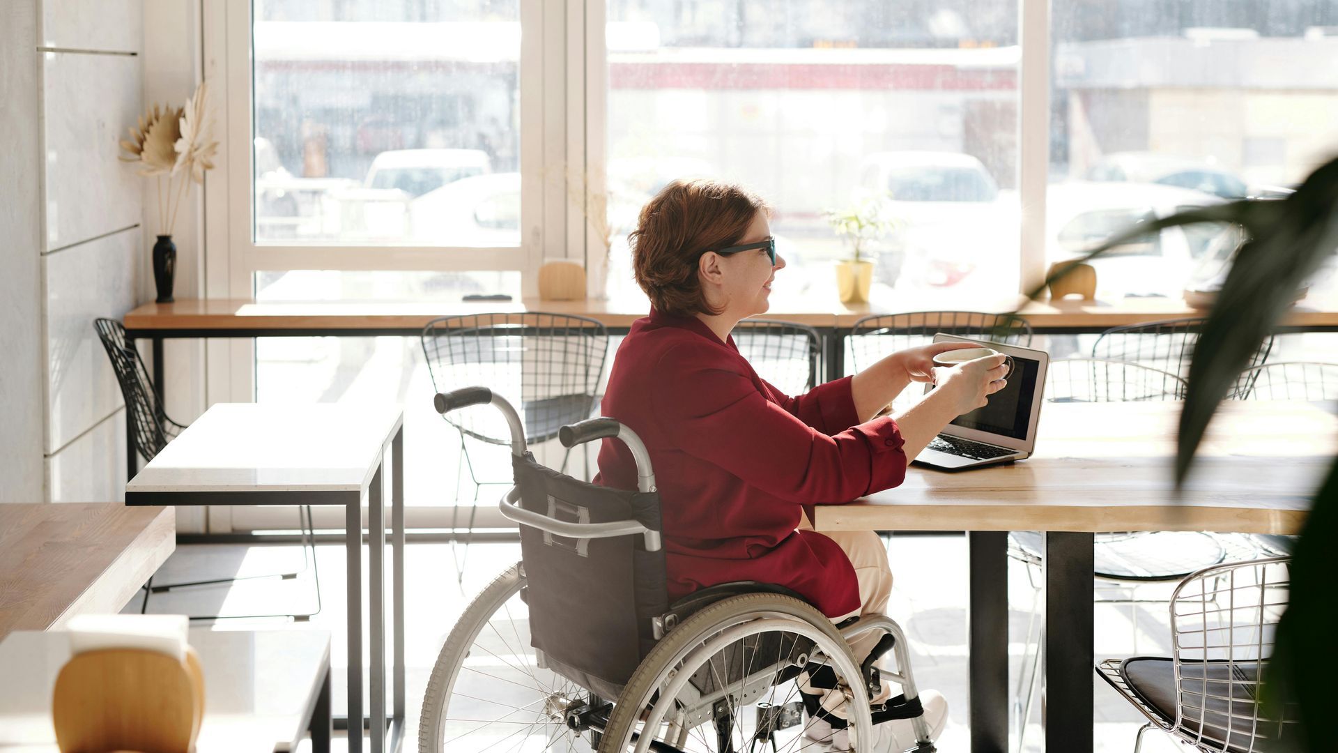 Woman in a wheelchair working on a laptop at a table near a window; red shirt, indoor setting.