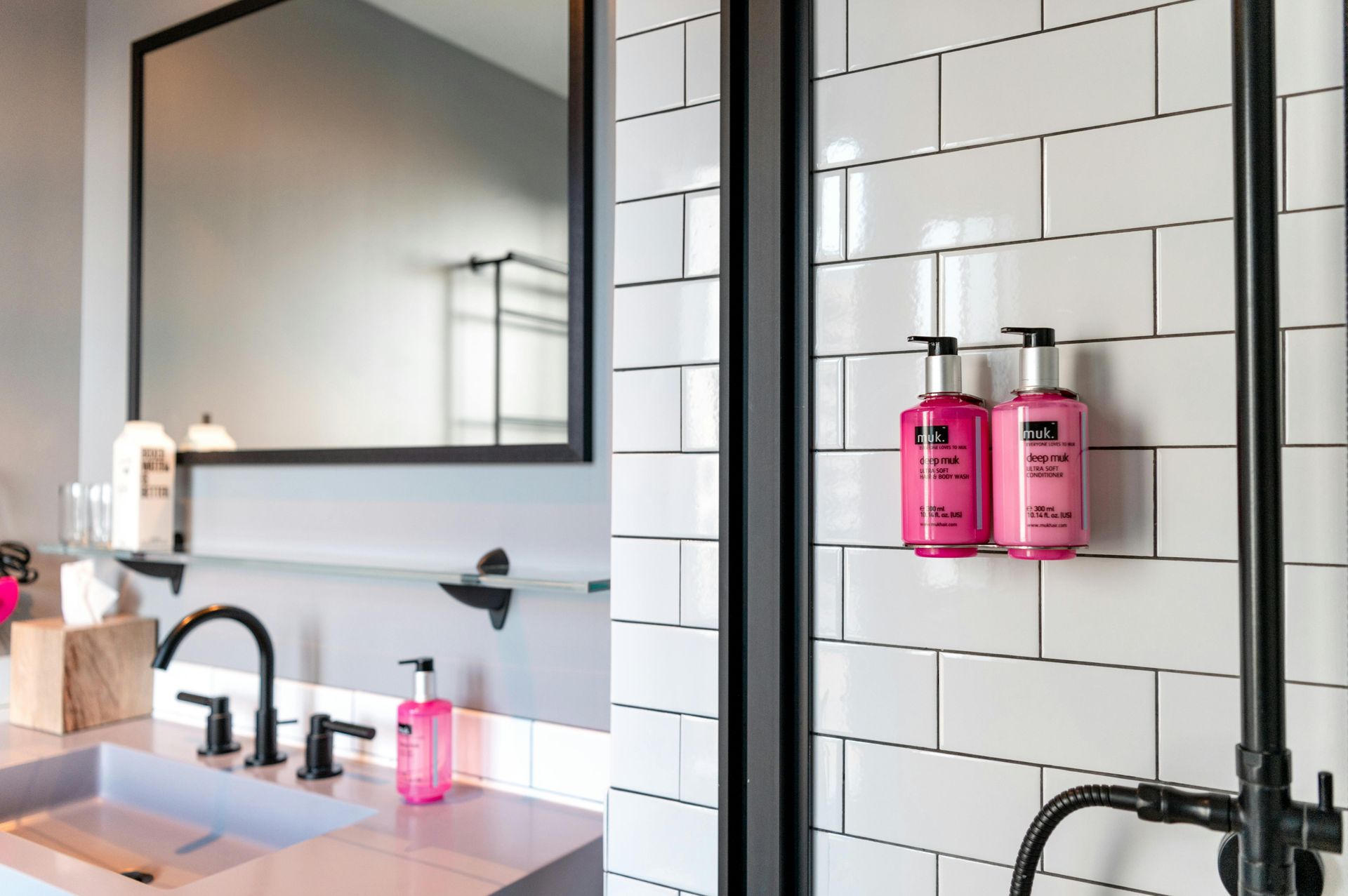 Modern bathroom with pink accents: shower bottles on white tile, black fixtures, and a large mirror.