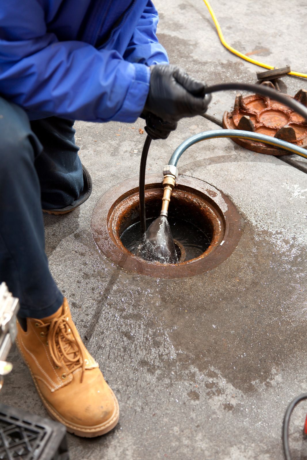 A person in a blue jacket and work boots cleans a sewer or drain using a high-pressure hose and a mechanical snake.
