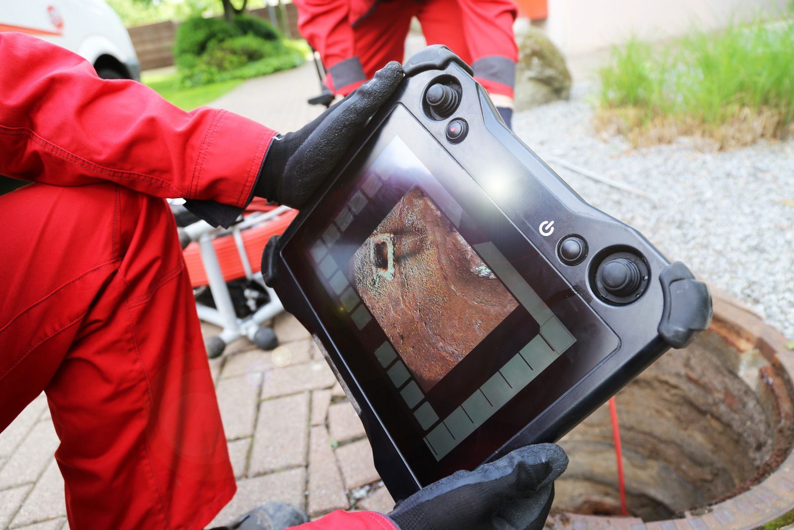 A technician in a red uniform holds a tablet displaying a live video feed of a pipe interior while performing an inspection.