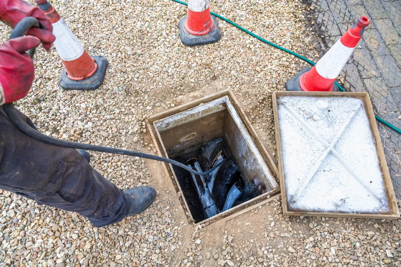 A worker in red gloves uses a high-pressure hose to clean an open drain in a gravel area marked by safety cones.