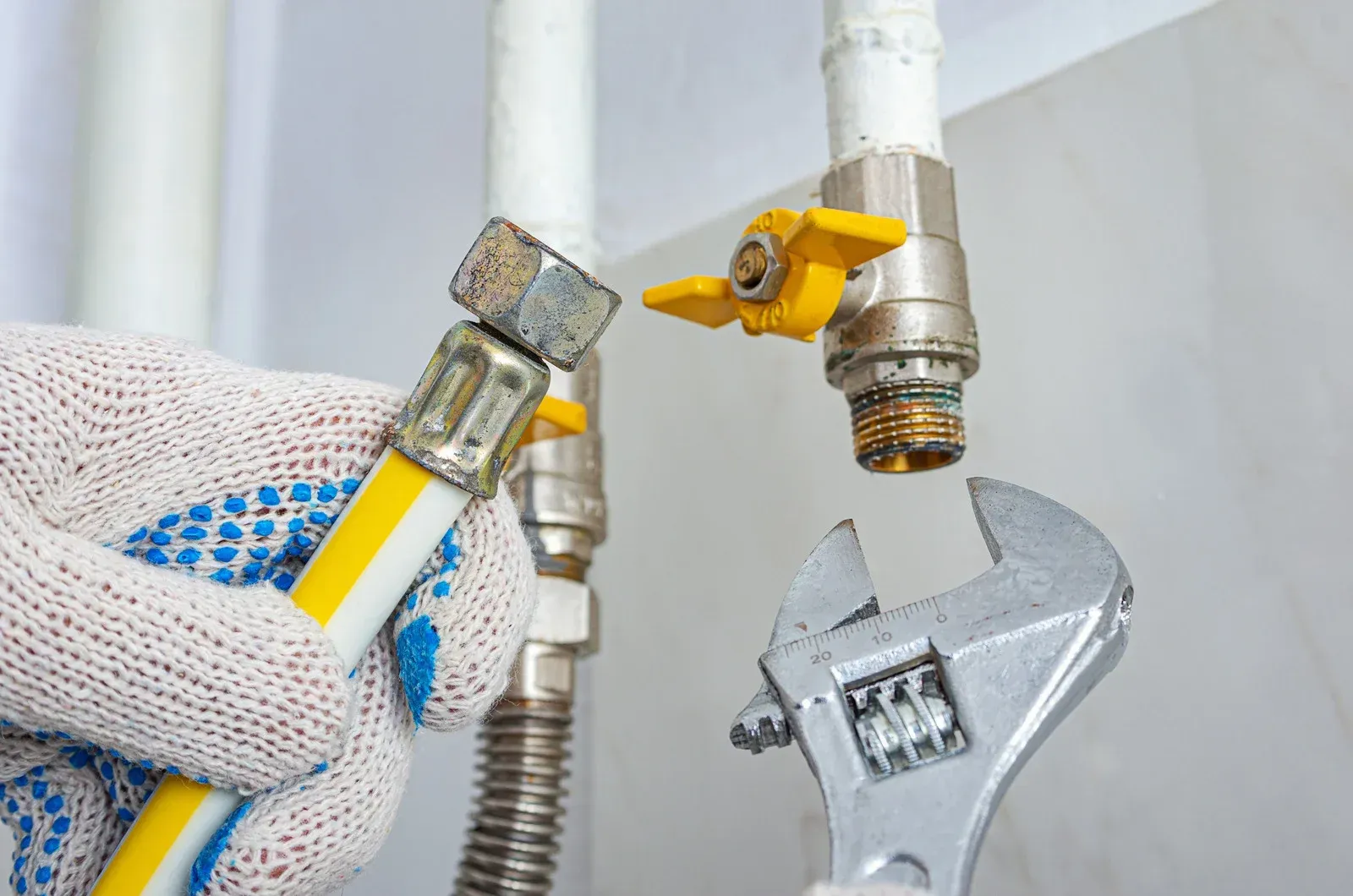 A hand in a protective glove holds a hose connector near a gas valve, with an adjustable wrench positioned nearby.