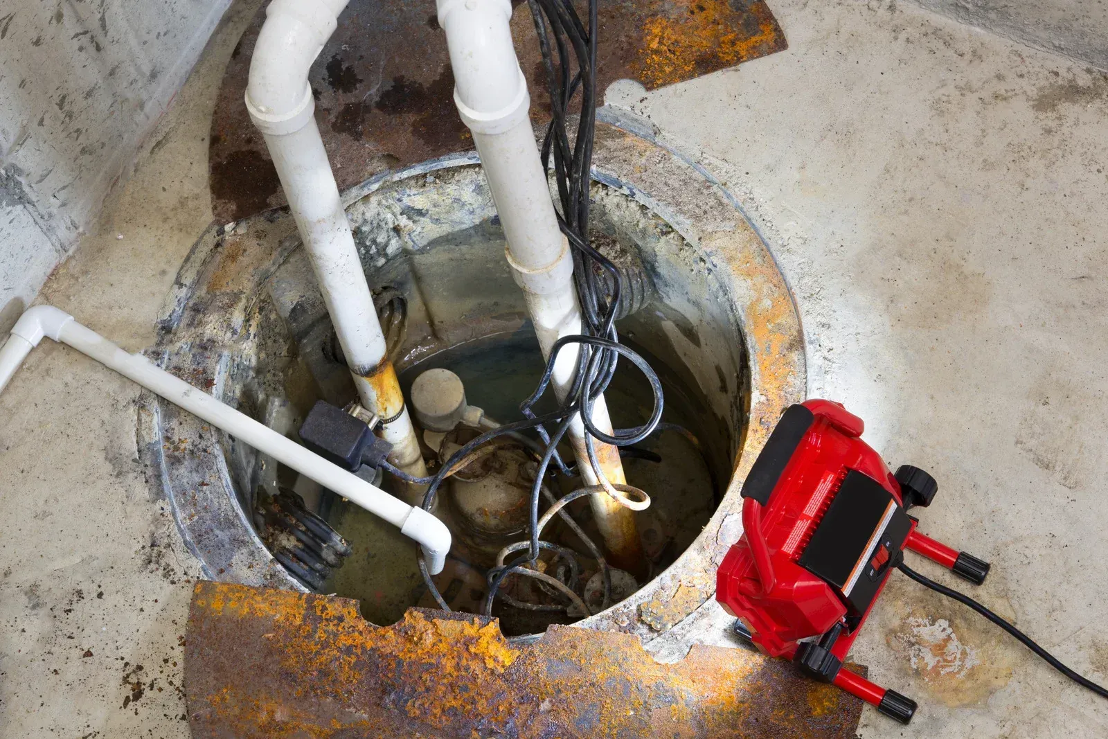 A sump pump with white PVC pipes sits in a concrete pit, illuminated by a red work light on the floor.