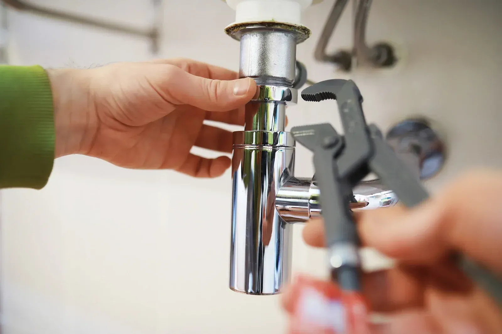 Close-up of hands using a plumbing wrench to tighten a chrome metal sink drain trap pipe.