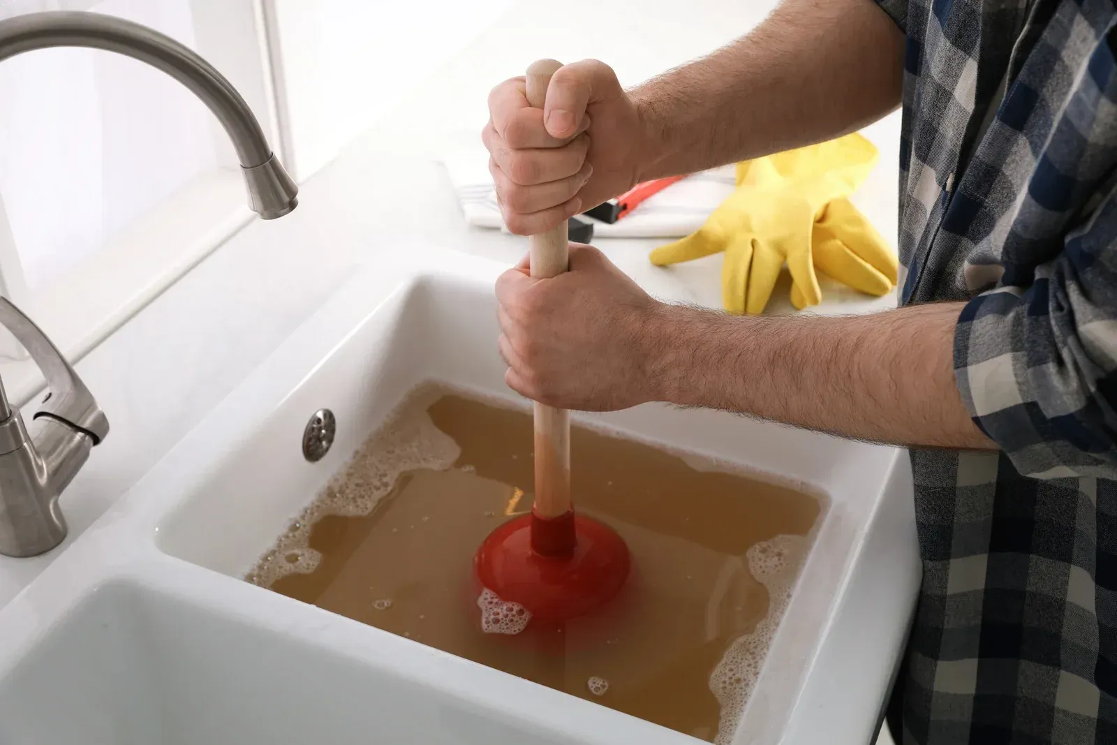 A person in a plaid shirt using a red plunger to clear a clogged, water-filled kitchen sink.