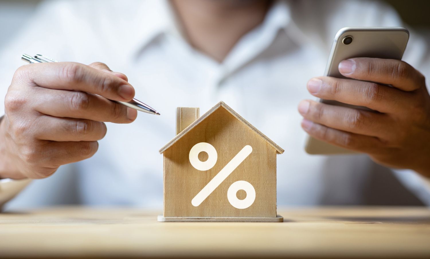 A person is holding a pen and a cell phone next to a wooden house with a percent sign on it.