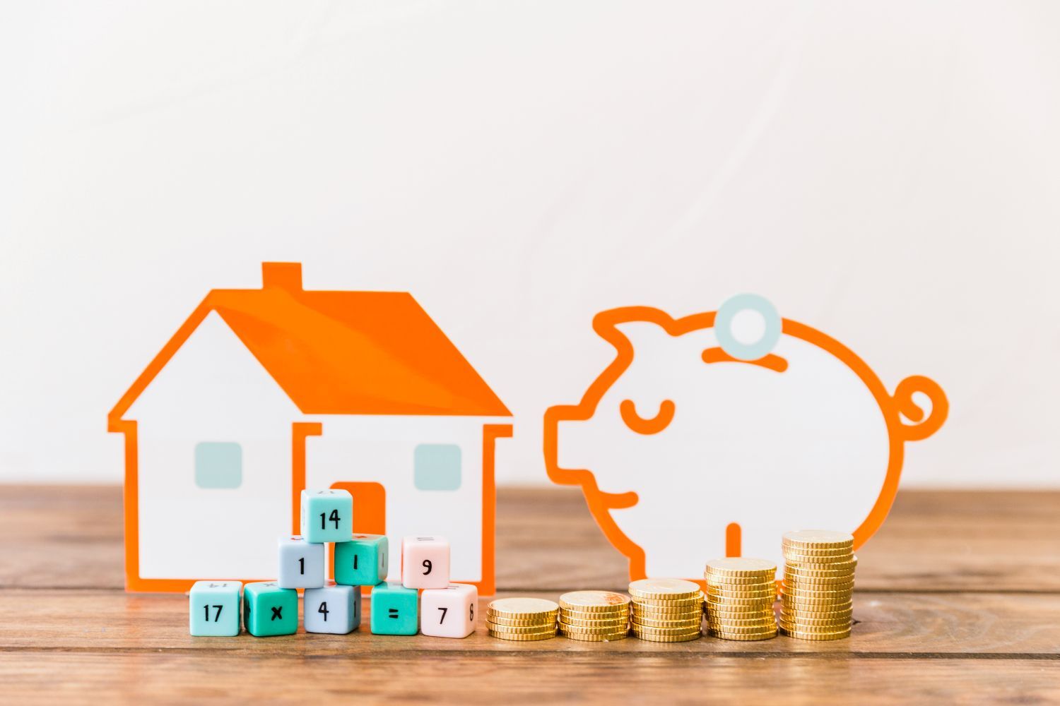 A piggy bank with a house and stacks of coins on a wooden table.