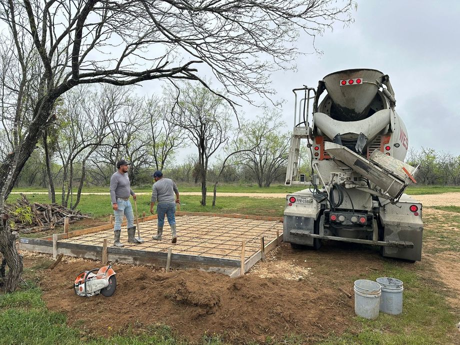 Two workers stand by a concrete mixer truck parked beside a wooden form filled with gravel for a foundation construction.