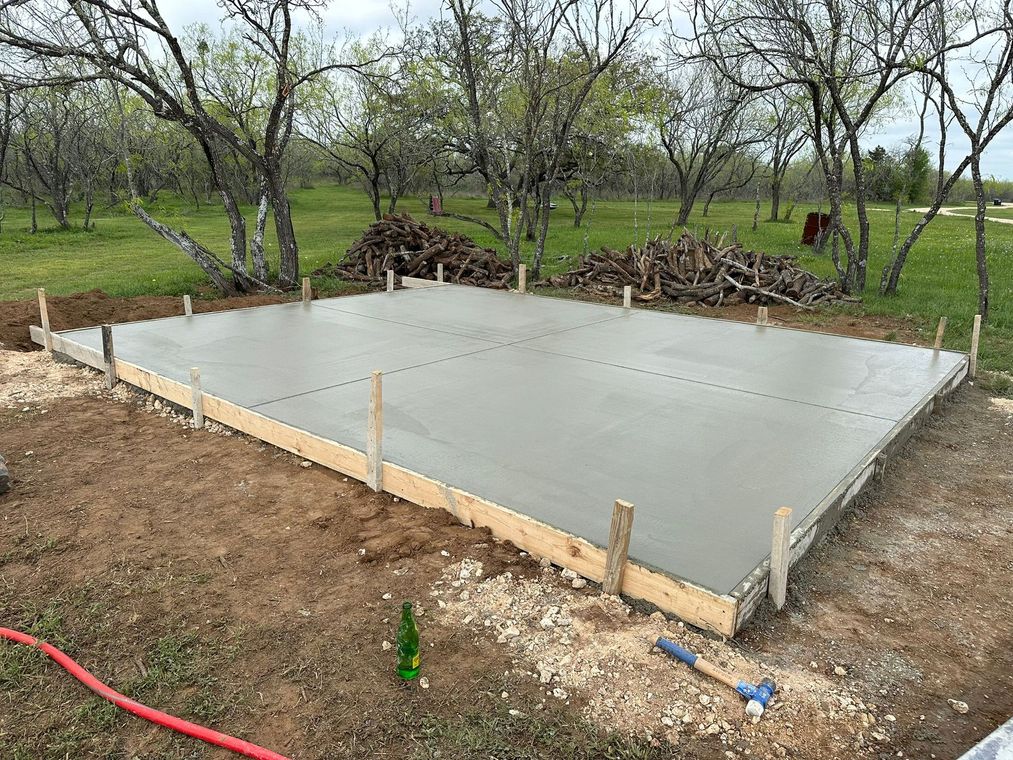 A newly poured rectangular concrete slab foundation in a grassy, wooded area, held by wooden formwork.