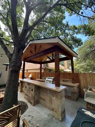 Outdoor kitchen with stone bar, built-in grill, and a wooden gazebo roof, set on a patio under a large tree.