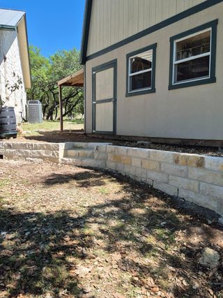 Tan shed with a dark-trimmed door and windows above a low, light-colored stone retaining wall with steps.