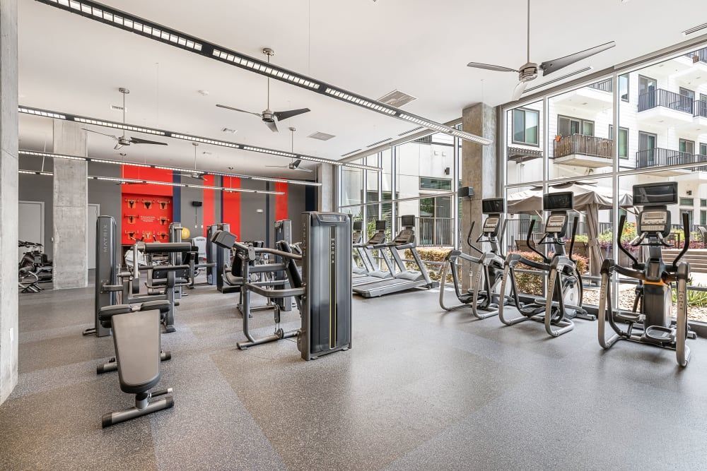 Modern gym interior with a variety of exercise equipment including treadmills, elliptical machines, and weight training stations. The space features large windows offering natural light, a bright red accent wall with mounted TVs, and a view of an outdoor patio area at Marq on Burnet in Austin, TX.